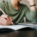 Image of person writing in notebook with pencil beside computer.